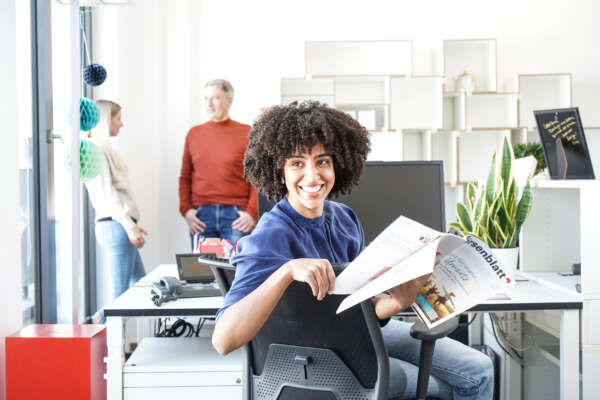 Frau sitzt an Arbeitsplatz im Büro mit einer Zeitschrift in der Hand.