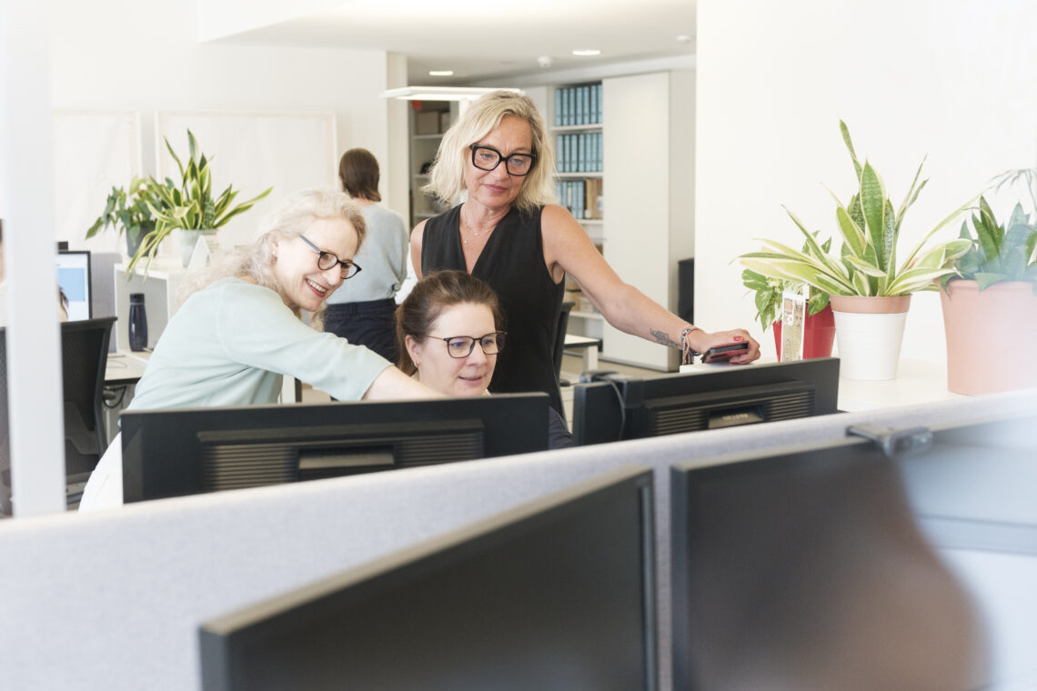 Eine Frau sitzt am Computer und zwei andere Frauen stehen rechts und links daneben und zeigen auf den Bildschirm.