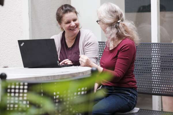 Zwei lachende Frauen, die sich anschauen und vor einem Laptop sitzen.