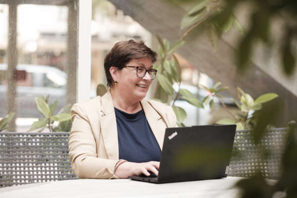 Eine lächelnde Frau sitzt an einem Tisch vor einem Laptop.