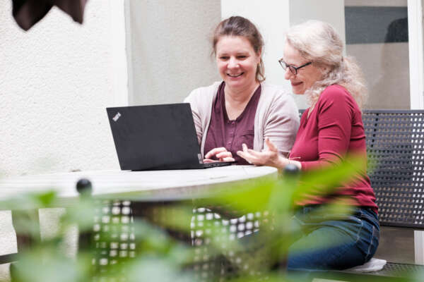 Zwei Frauen sitzen draußen an einem Tisch und gucken auf einen Laptop.