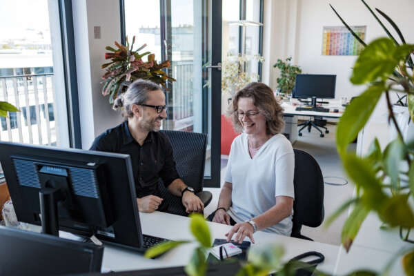 Kollegin und Kollege im begrünten Büro im Gespräch vor PC