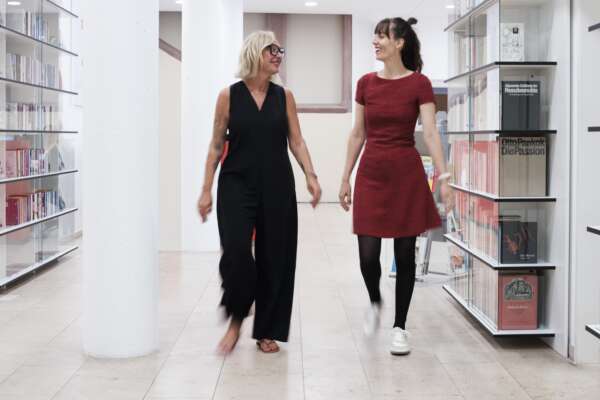 Zwei Frauen laufen durch einen Flur mit Bücherregalen an der Wand.