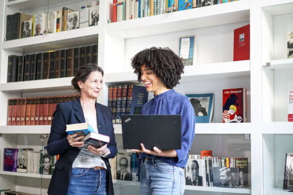 Zwei Frauen stehen vor einem Bücherregal und schauen auf Bücher und Laptop.
