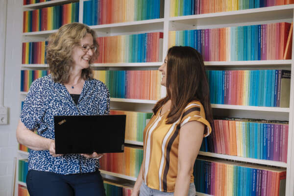 Zwei Frauen stehen vor einem Bücherregal, worin die Bücher nach einem Regenbogen sortiert sind.
