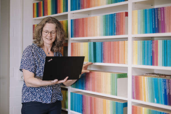Frau steht mit Laptop in der Hand neben einem Bücherregal.