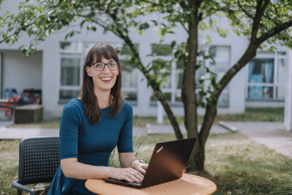 Eine Frau sitzt an einem Tisch im Freien vor einem Laptop und lächelt in die Kamera.