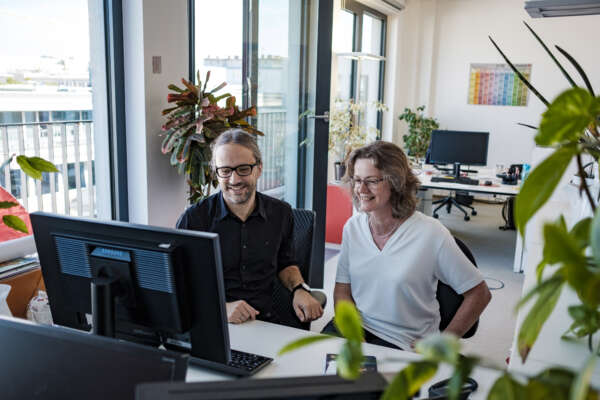Ein Mann (links) und eine Frau (rechts) sitzen gemeinsam an einem Schreibtisch und schauen auf den Bildschirm.