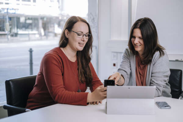 Zwei Frauen sitzen an einem Tisch und schauen auf ein stehendes Tablet.