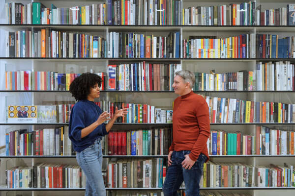 Frau (links) und Mann (rechts) stehen vor einem Bücherregal und unterhalten sich.